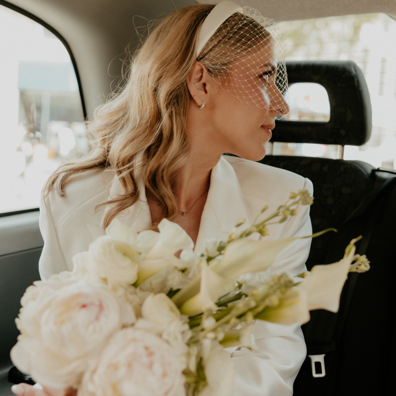 Bride wears a birdcage veil with a slim ivory padded headband and an ivory blazer mini dress with a bouquet of call lillies and peonies