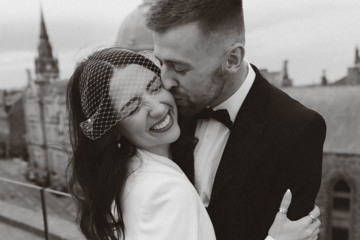bride wears luxury birdcage veil for her chic city hall wedding in an embrace with her tuxedo-wearing groom who is kissing her while she smiles happily