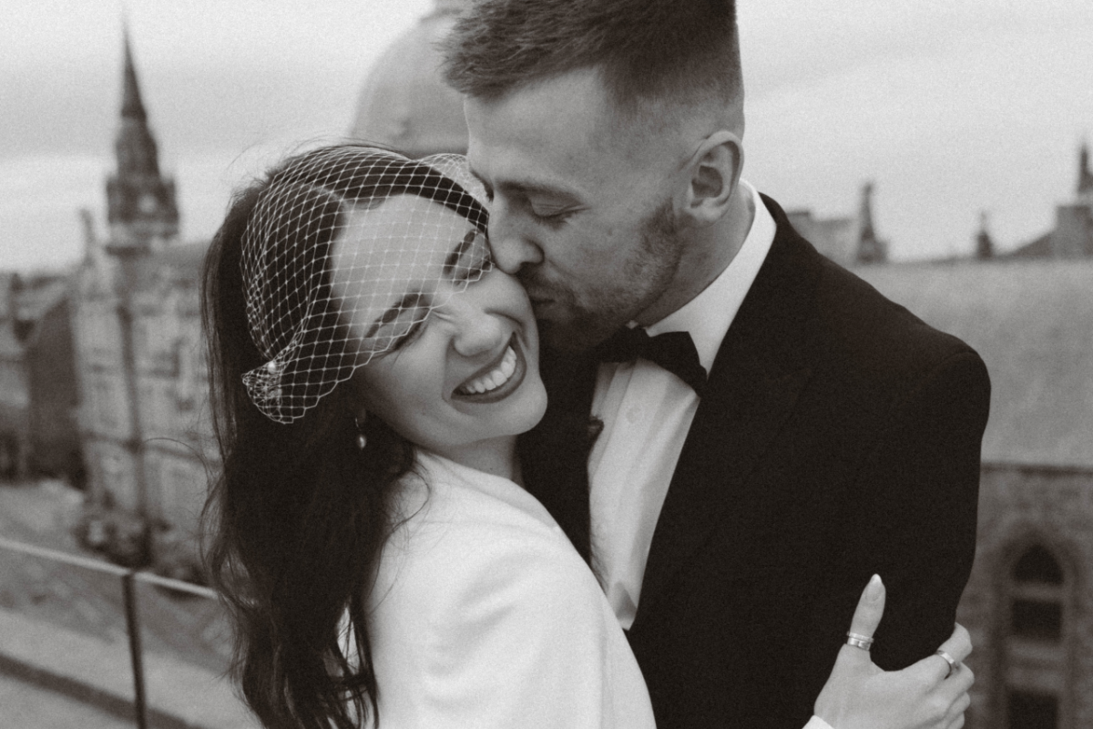 bride wears luxury birdcage veil for her chic city hall wedding in an embrace with her tuxedo-wearing groom who is kissing her while she smiles happily