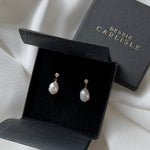 A pair of Debbie Carlisle pearl and crystal stud drop earrings displayed in an open black presentation box, with the branded closed box behind it on draped ivory fabric.