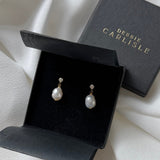 A pair of Debbie Carlisle pearl and crystal stud drop earrings displayed in an open black presentation box, with the branded closed box behind it on draped ivory fabric.