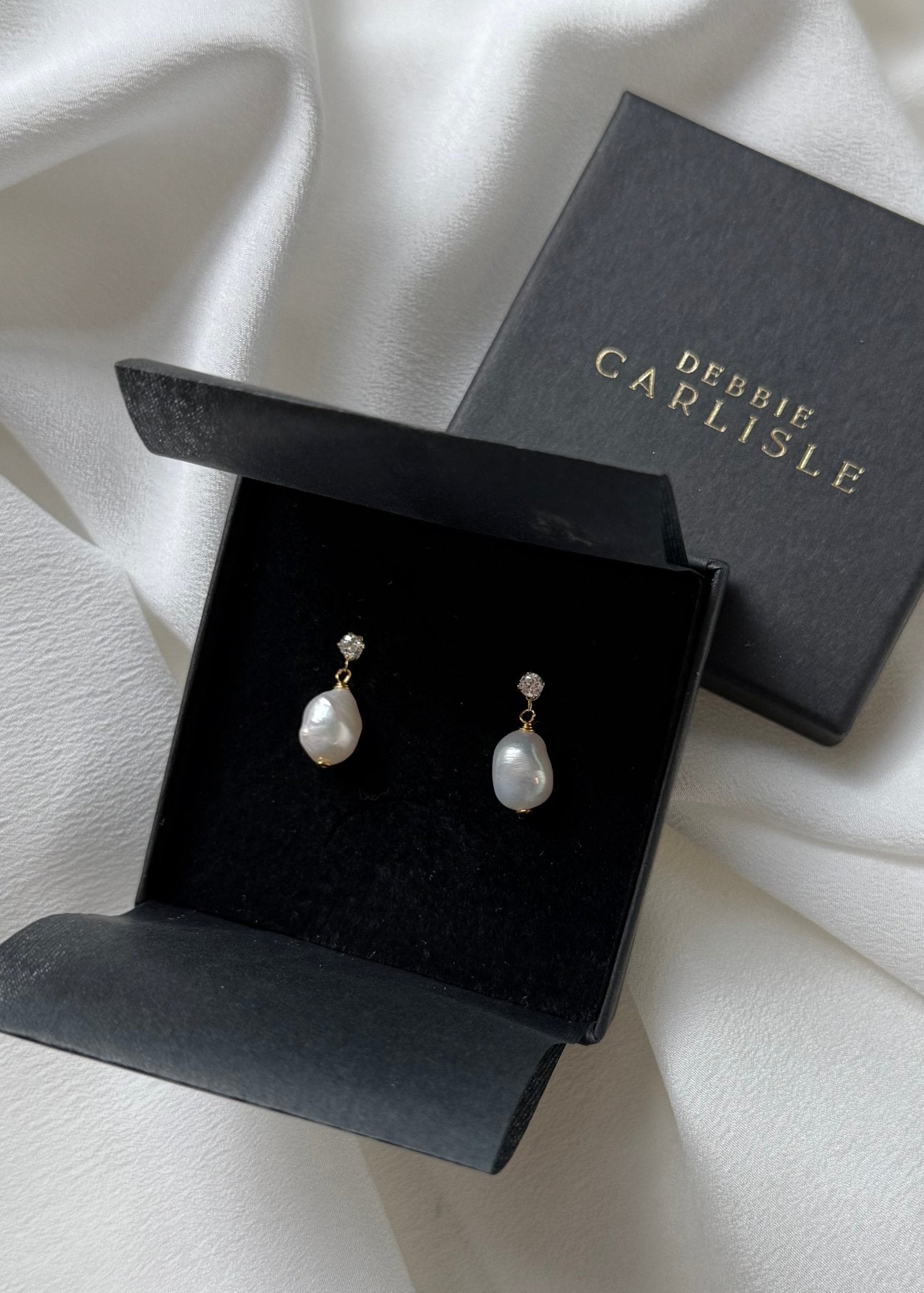 A pair of Debbie Carlisle pearl and crystal stud drop earrings displayed in an open black presentation box, with the branded closed box behind it on draped ivory fabric.