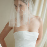 Woman wearing a white wedding dress with a bow-embellished veil against a neutral background