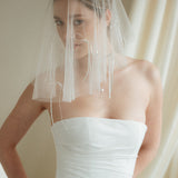 Woman wearing a white wedding dress with a bow-embellished veil against a neutral background
