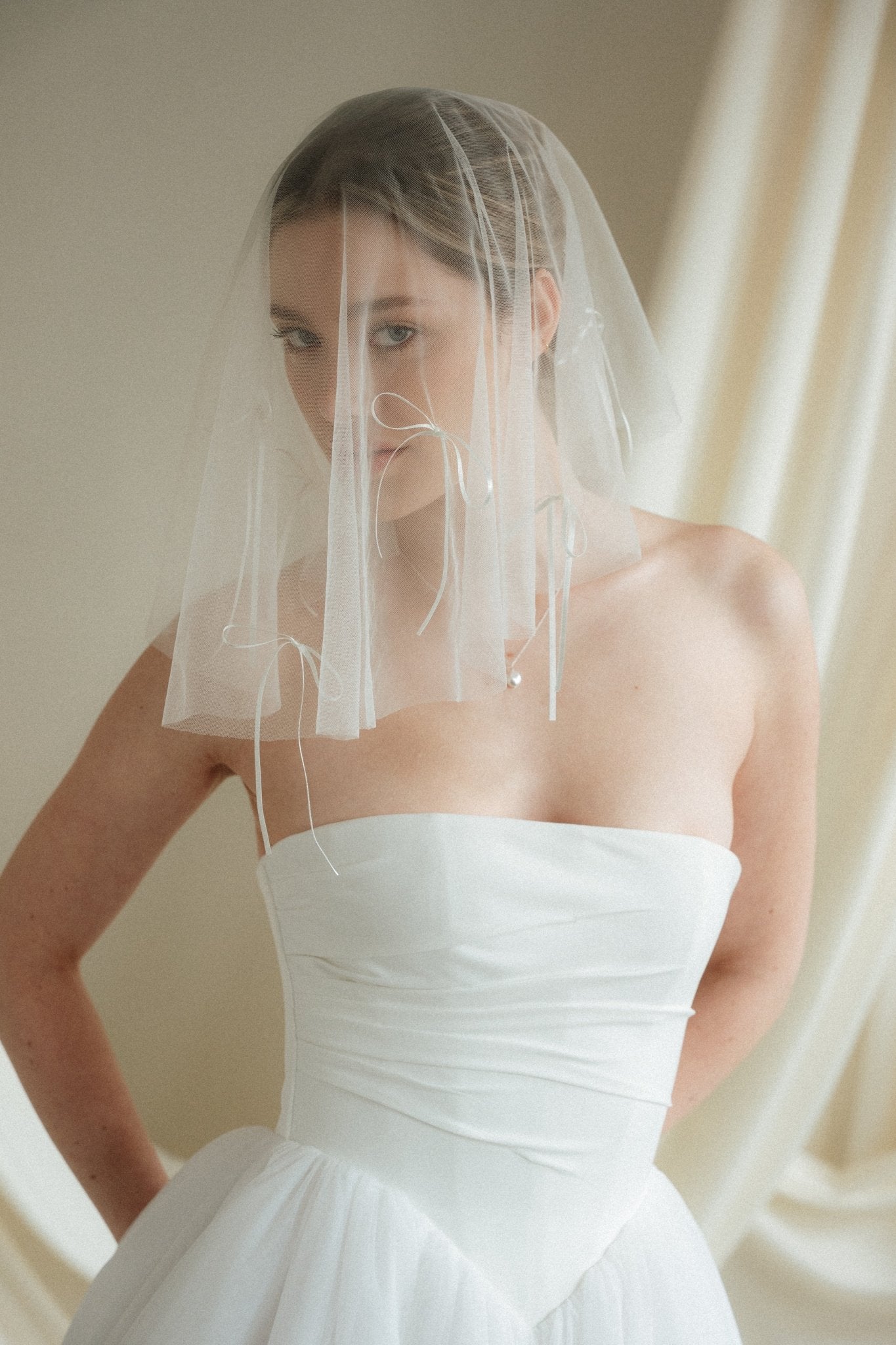 Woman wearing a white wedding dress with a bow-embellished veil against a neutral background