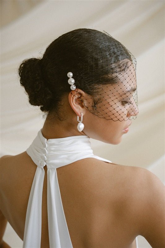 Black bride wears a black mask style Birdcage Veil with a pearl comb consisting of three pearls and a pair of statement pearl earrings and a halterneck wedding dress.