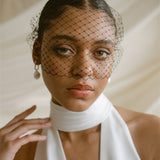 Black bride wears a black mask style Birdcage Veil with statement pearl earrings and a halterneck wedding dress.