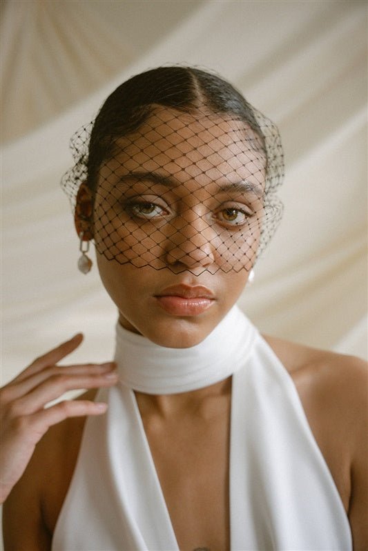 Black bride wears a black mask style Birdcage Veil with statement pearl earrings and a halterneck wedding dress.