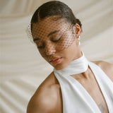 Black bride wears a black mask style Birdcage Veil with statement pearl earrings and a halterneck wedding dress.