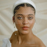 A black bride wears a full gathered birdcage veil with an ivory satin padded headband and statement long drop baroque pearl earrings