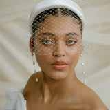 A black bride wears a full gathered birdcage veil with an ivory satin padded headband and statement long drop baroque pearl earrings