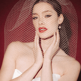 Bride wearing a white dress and long blusher length birdcage veil against a red background