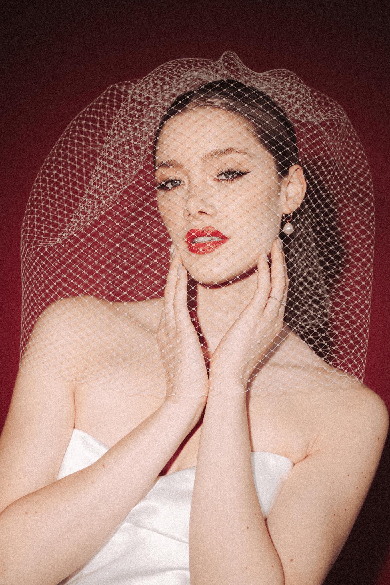 Bride wearing a white dress and long blusher length birdcage veil against a red background