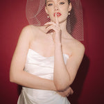 Bride wearing a white dress and long blusher length birdcage veil against a red background