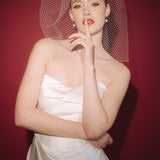 Bride wearing a white dress and long blusher length birdcage veil against a red background