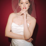 Bride wearing a white dress and long blusher length birdcage veil against a red background