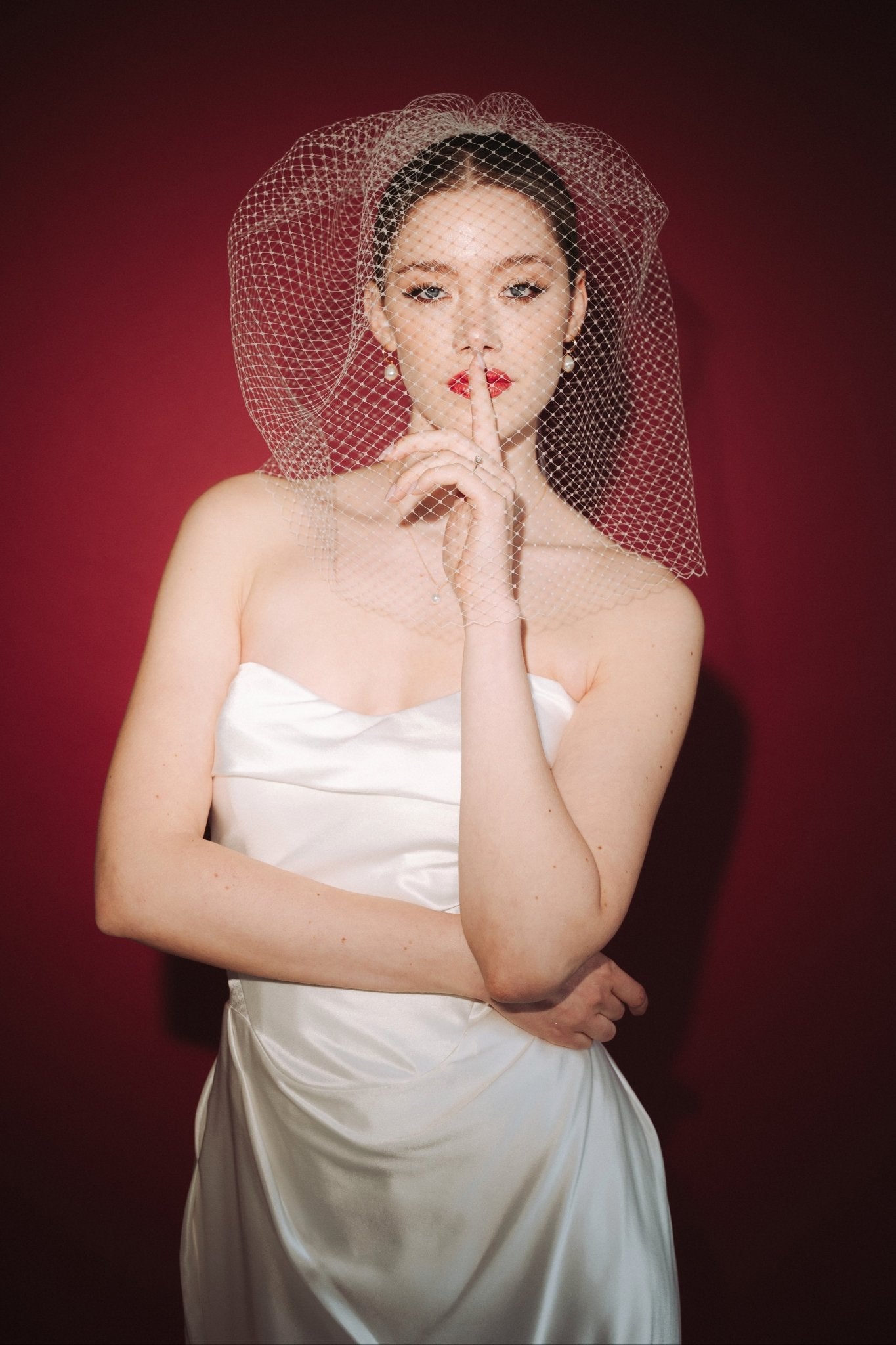 Bride wearing a white dress and long blusher length birdcage veil against a red background