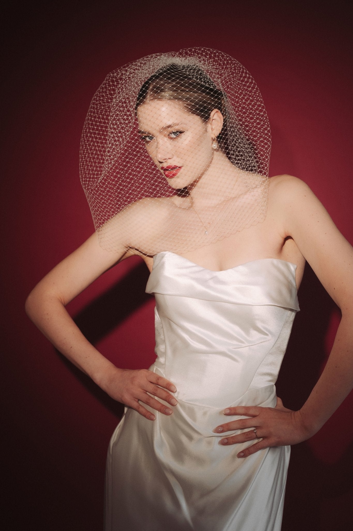 Bride wearing a white dress and long blusher length birdcage veil against a red background