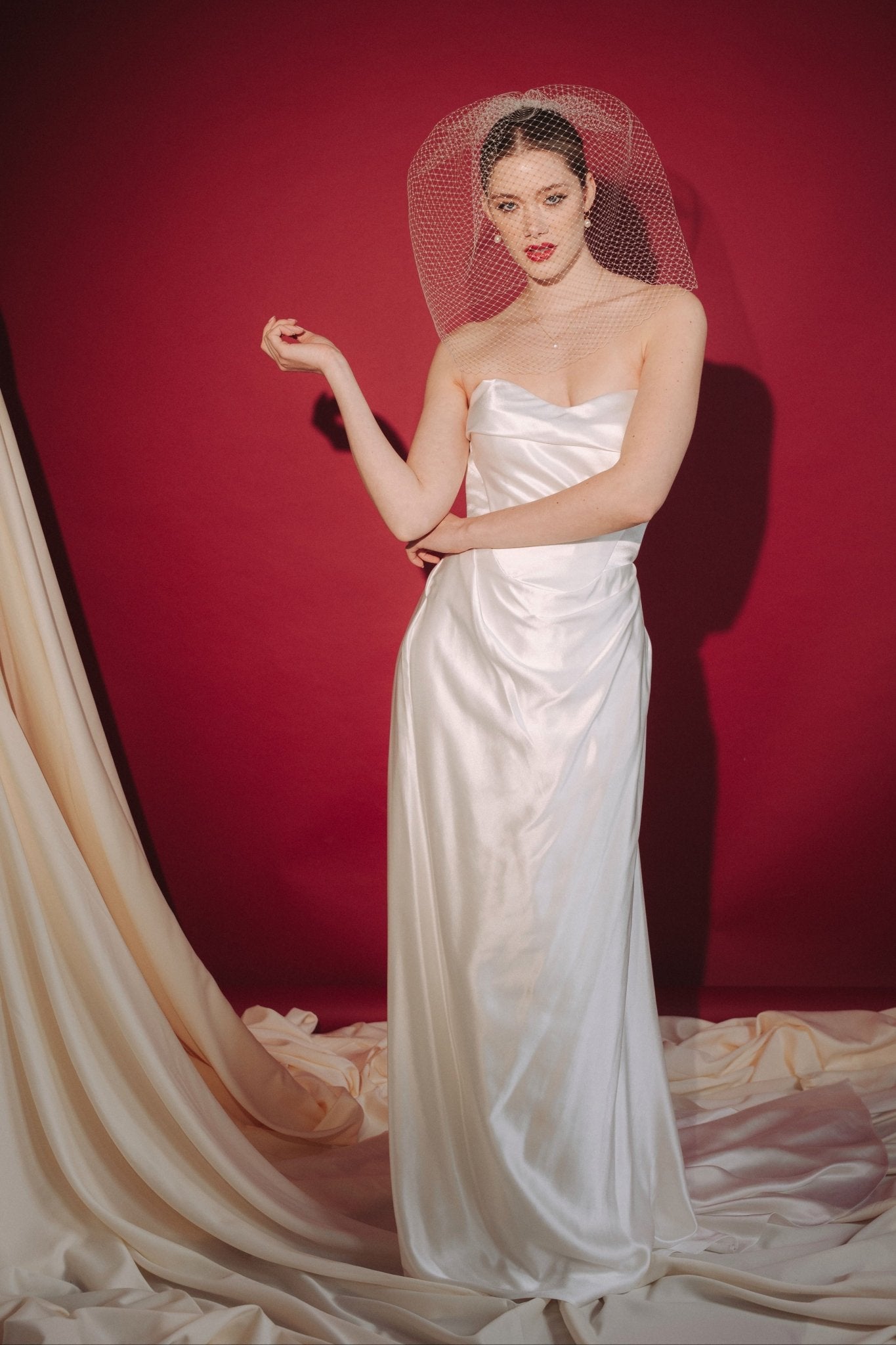 Bride wearing a white dress and long blusher length birdcage veil against a red background
