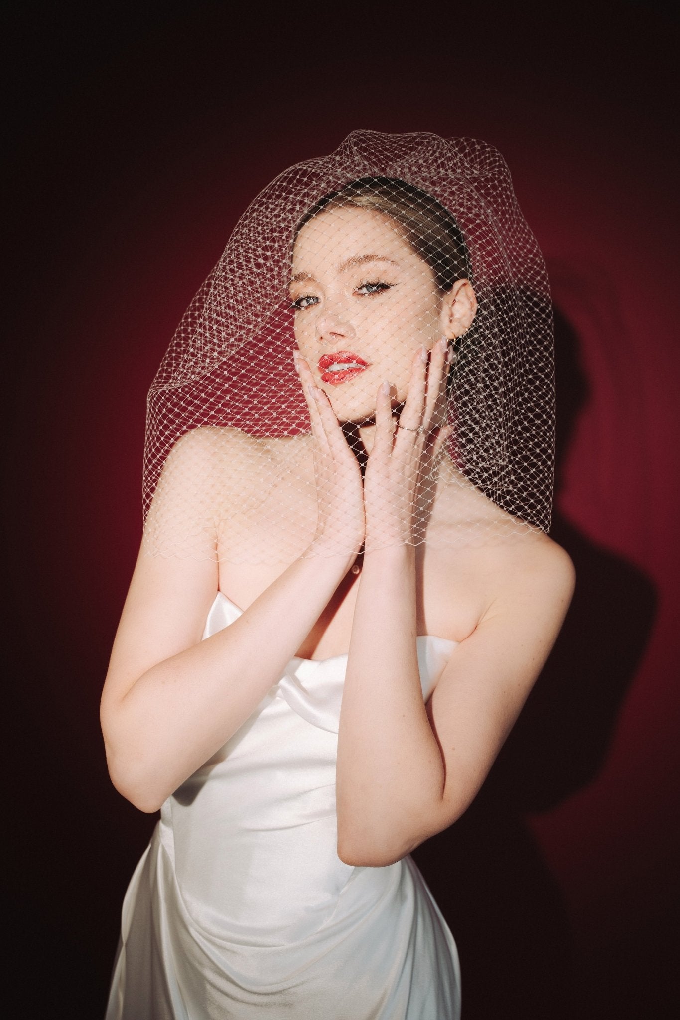 Bride wearing a white dress and long blusher length birdcage veil against a red background
