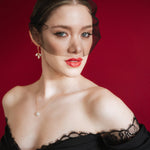 Bride wearing a black off-shoulder dress with lace details, a gold baroque coin vegan pearl necklace, gold crystal drop earrings and a black tulle mask veil micro veil against a red background