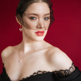 Bride wearing a black off-shoulder dress with lace details, a gold baroque coin vegan pearl necklace, gold crystal drop earrings and a black tulle mask veil micro veil against a red background
