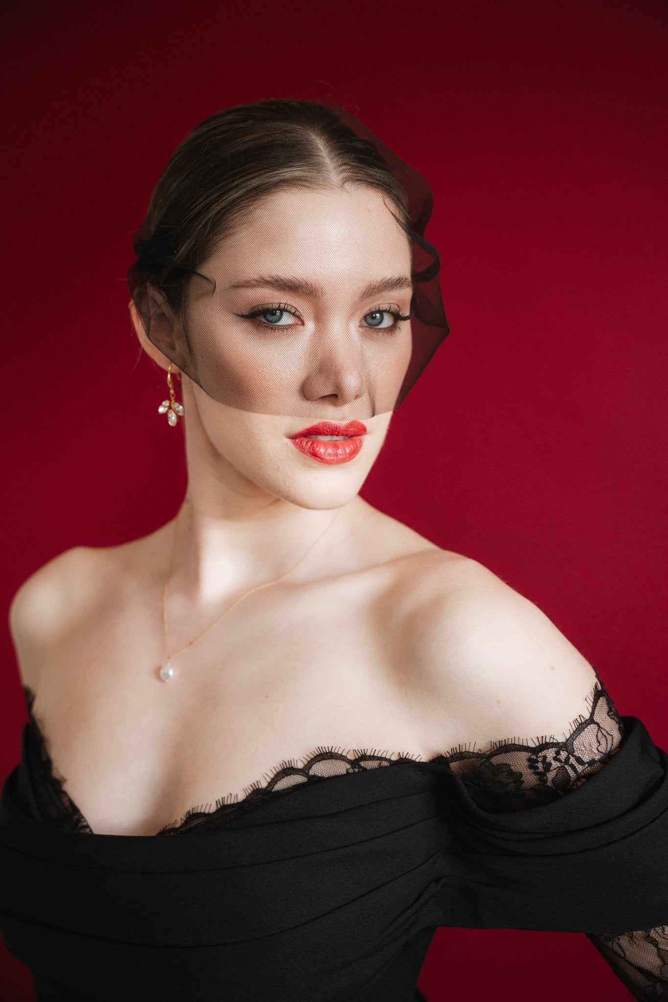 Bride wearing a black off-shoulder dress with lace details, a gold baroque coin vegan pearl necklace, gold crystal drop earrings and a black tulle mask veil micro veil against a red background