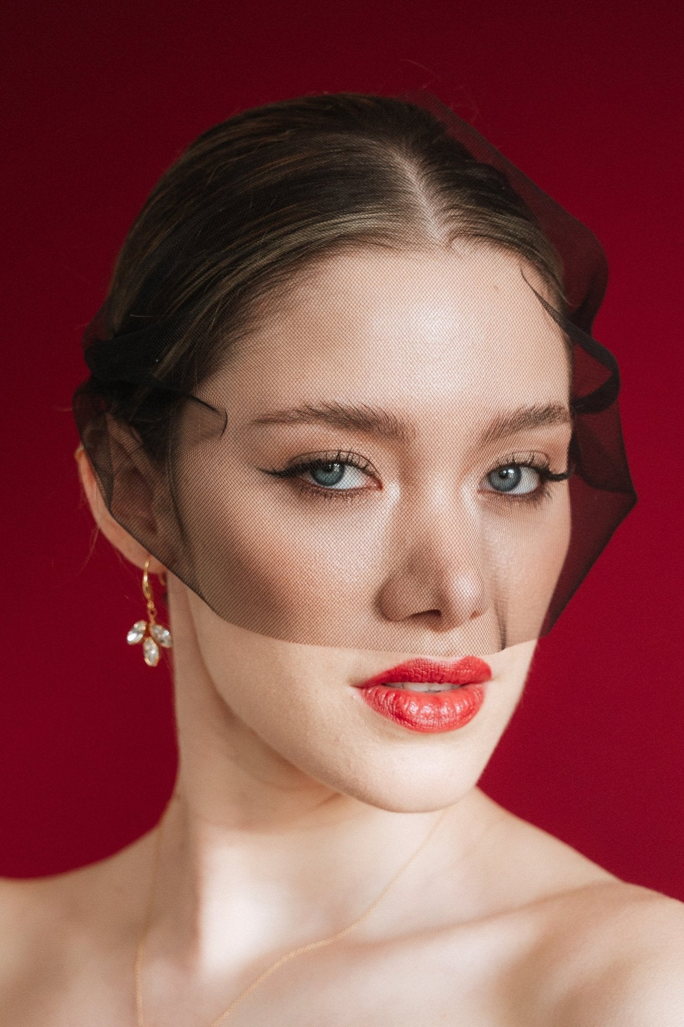 Bride wearing gold crystal drop earrings and a black tulle mask veil against a red background