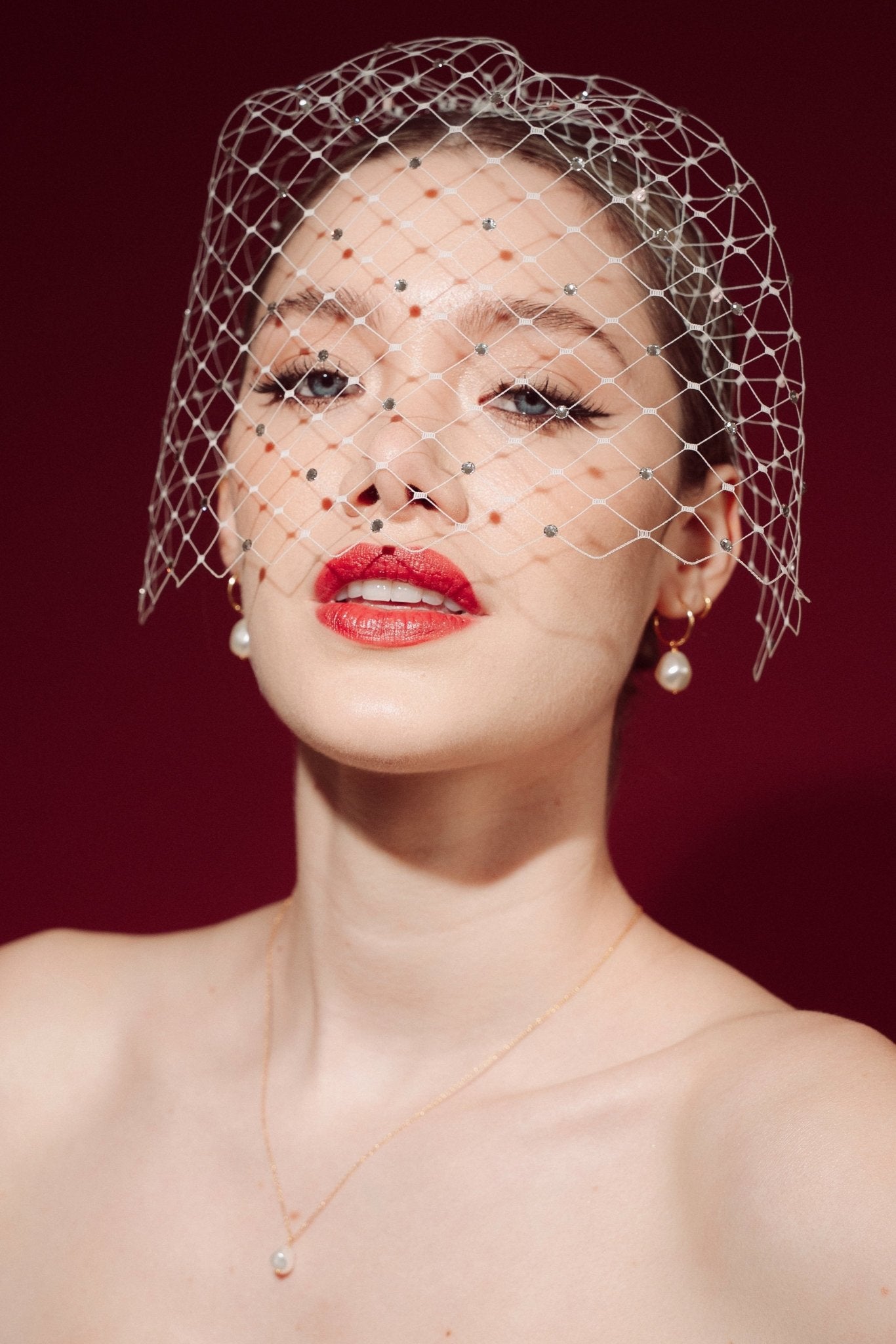 Woman wearing a white strapless dress and crystal birdcage veil against a red background