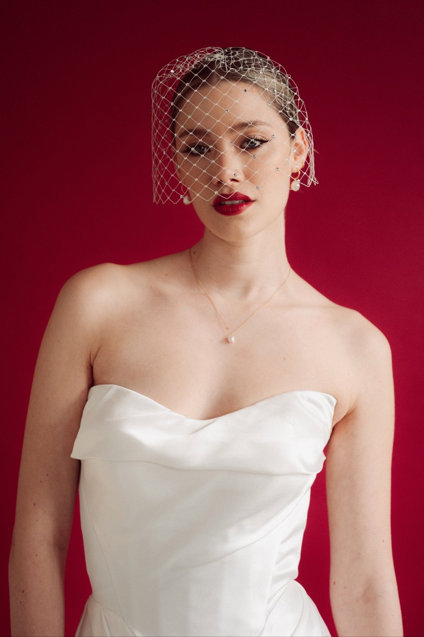 Woman wearing a white strapless dress and crystal birdcage veil against a red background