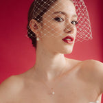 Woman wearing a white strapless dress and crystal birdcage veil against a red background  with baroque pearl hoop earrings and matching necklace