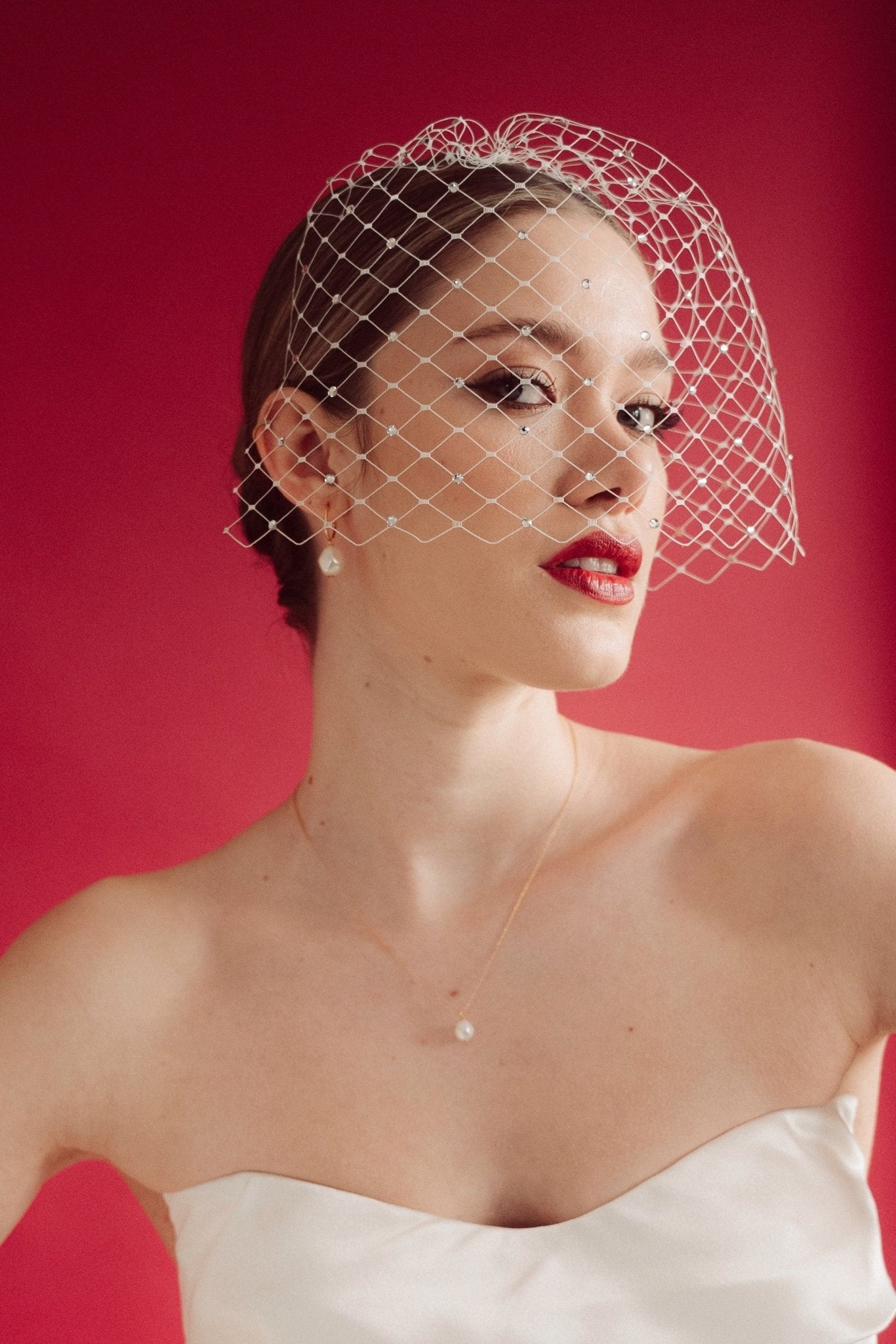 Woman wearing a white strapless dress and crystal birdcage veil against a red background  with baroque pearl hoop earrings and matching necklace