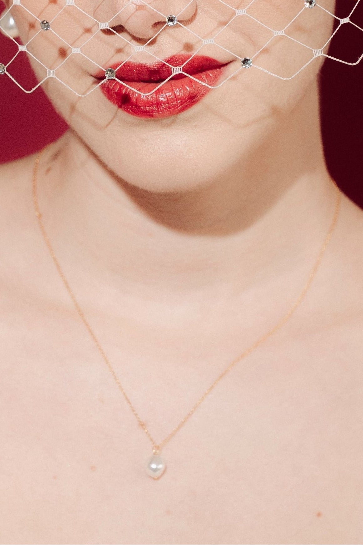 Woman wearing a white strapless dress and crystal birdcage veil against a red background with a baroque pearl necklace and matching earrings