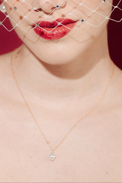 Woman wearing a white strapless dress and crystal birdcage veil against a red background with a baroque pearl necklace and matching earrings