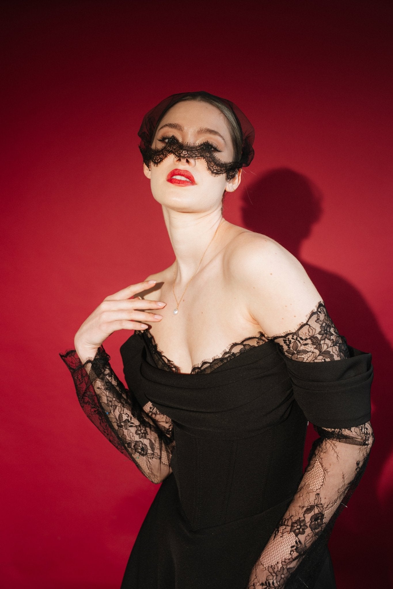 Woman in a black lace dress against a red background wears a black mask style micro veil with a lace edging