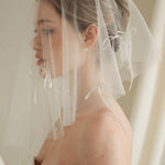 Woman wearing a white wedding dress with a bow-embellished veil against a neutral background