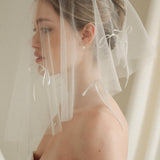 Woman wearing a white wedding dress with a bow-embellished veil against a neutral background