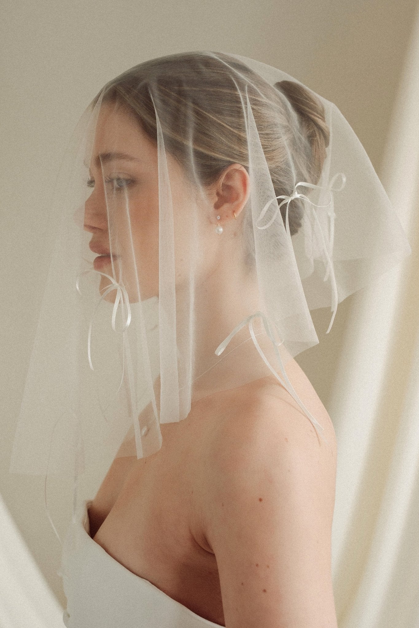 Woman wearing a white wedding dress with a bow-embellished veil against a neutral background