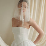 Woman wearing a white wedding dress with a bow-embellished veil against a neutral background