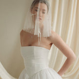 Woman wearing a white wedding dress with a bow-embellished veil against a neutral background