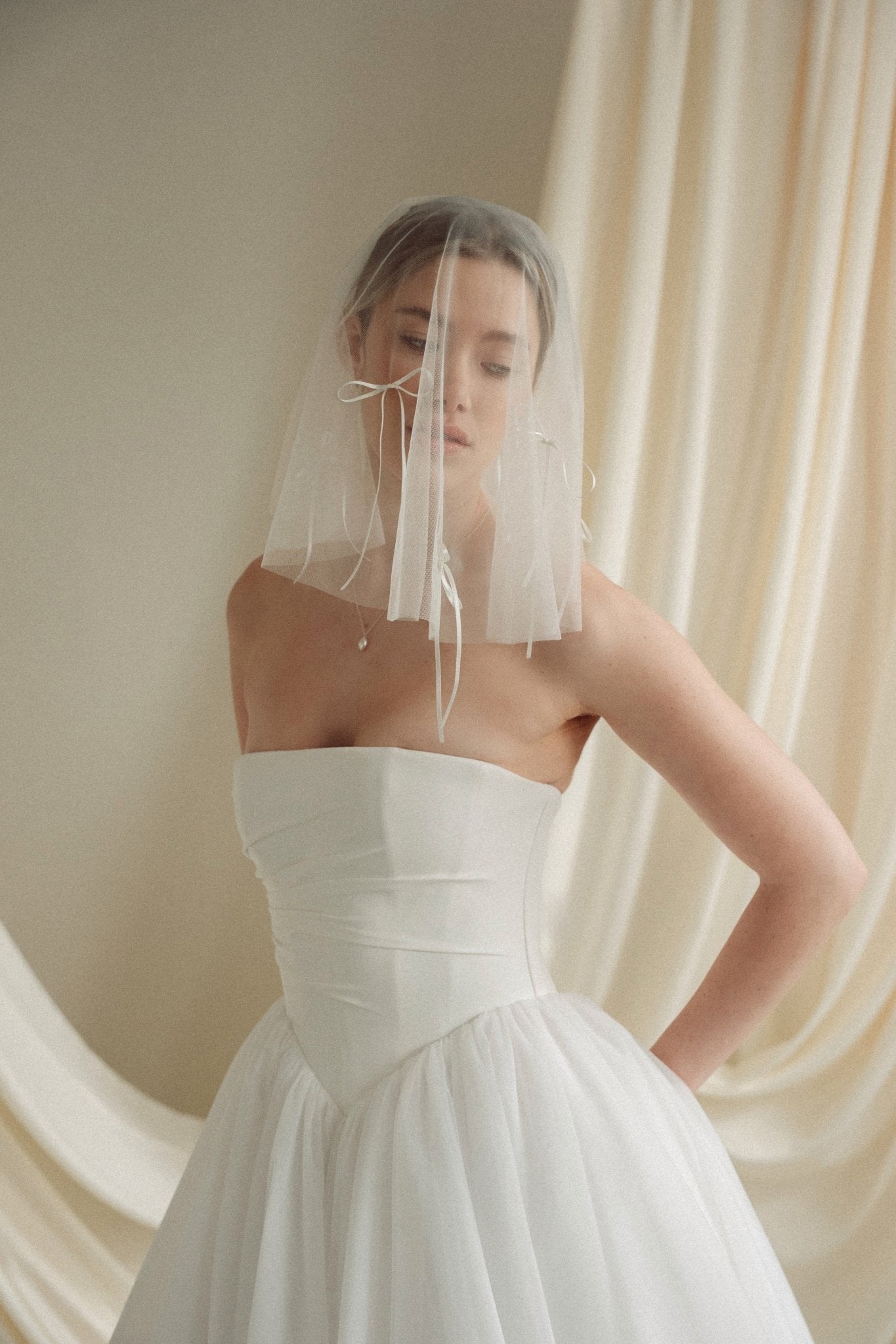 Woman wearing a white wedding dress with a bow-embellished veil against a neutral background