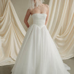 Woman wearing a white wedding dress with a bow-embellished veil against a neutral background