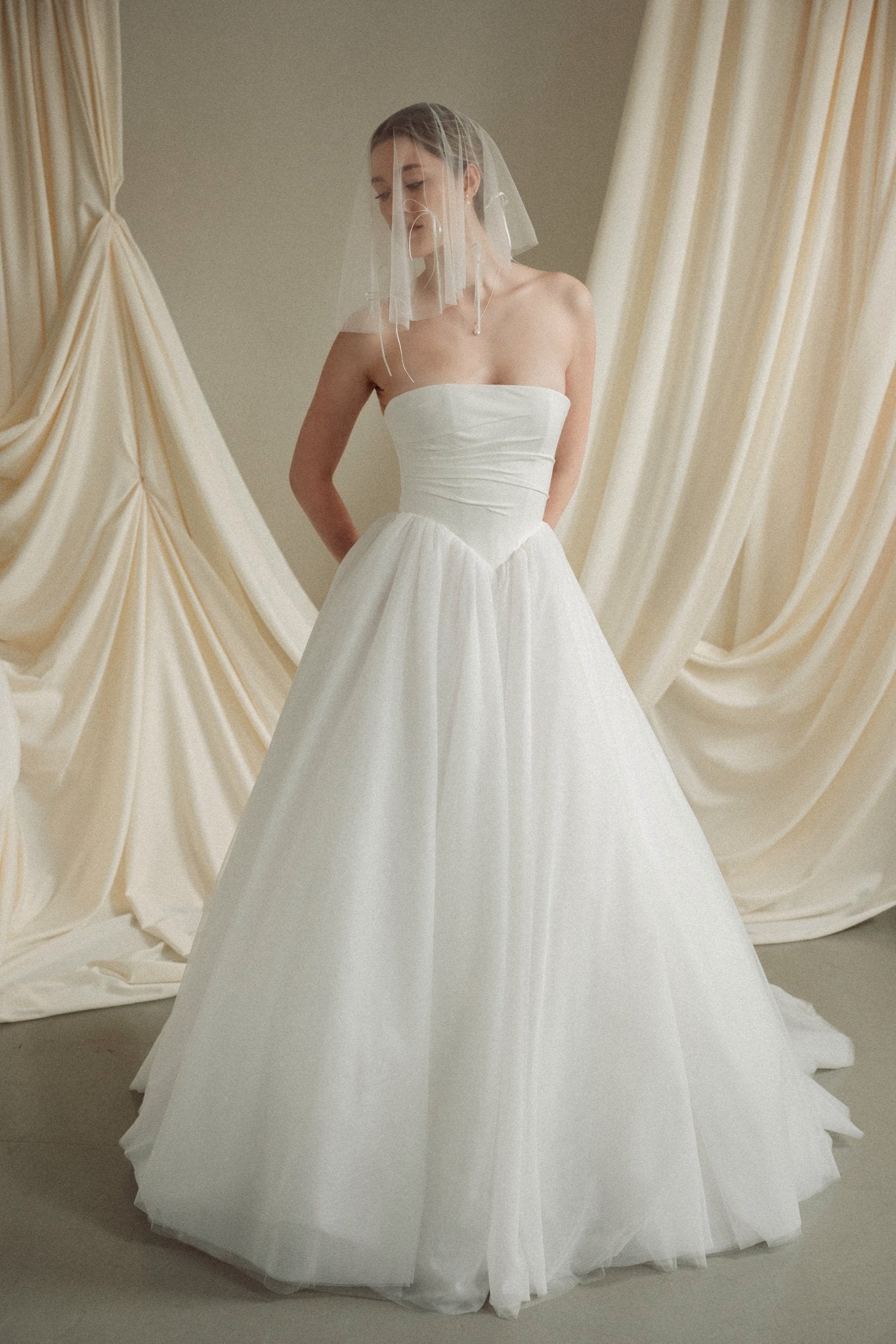 Woman wearing a white wedding dress with a bow-embellished veil against a neutral background