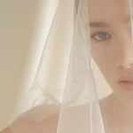 Woman wearing a white wedding dress with a bow-embellished veil against a neutral background