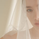 Woman wearing a white wedding dress with a bow-embellished veil against a neutral background