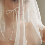Woman wearing a white wedding dress with a bow-embellished veil against a neutral background