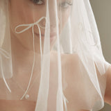 Woman wearing a white wedding dress with a bow-embellished veil against a neutral background