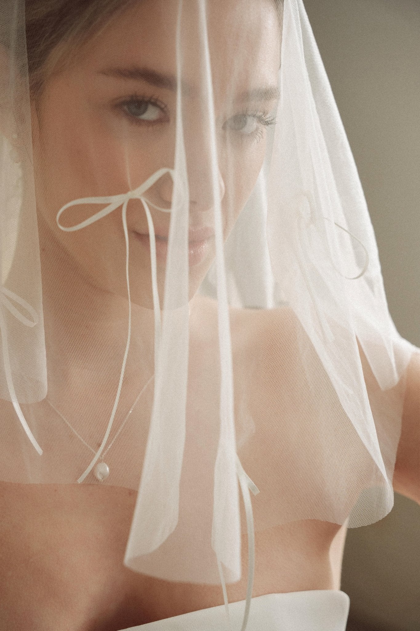 Woman wearing a white wedding dress with a bow-embellished veil against a neutral background
