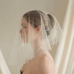 Woman wearing a white wedding dress with a bow-embellished veil against a neutral background