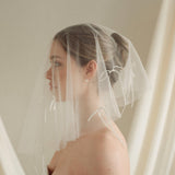 Woman wearing a white wedding dress with a bow-embellished veil against a neutral background